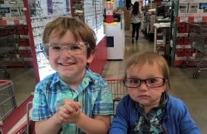 two kids sitting in a costco shopping cart, wearing glasses