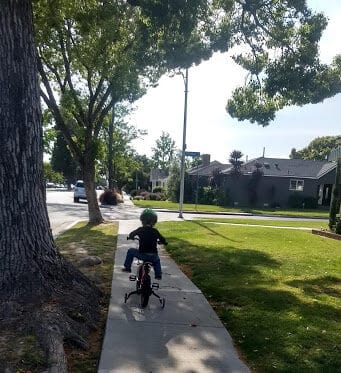 Toddler on a bicycle with training wheels outside on the sidewalk.