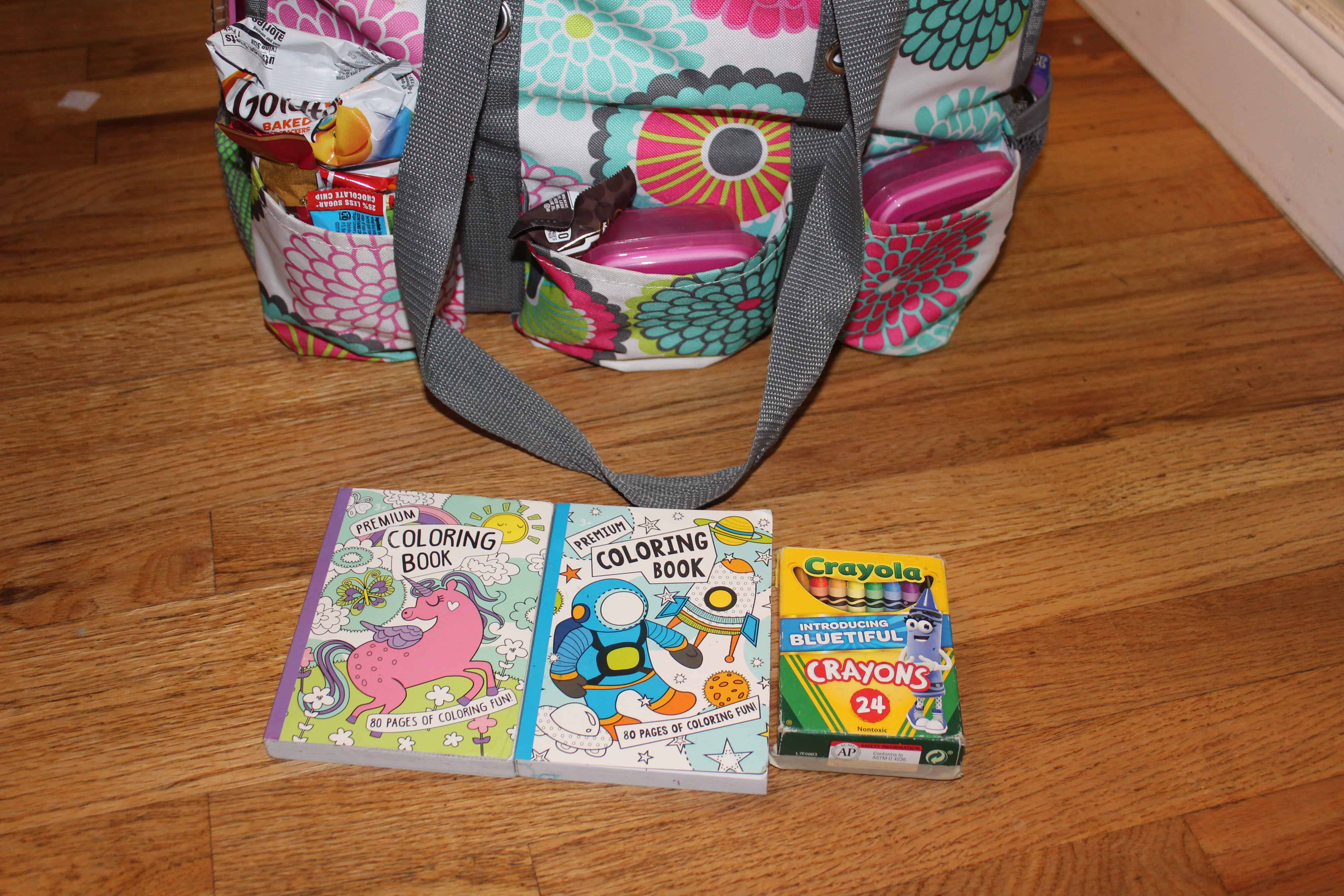 Coloring books and a pack of 24 crayons in front of a travel bag.