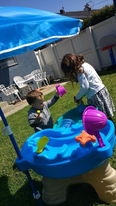 A toddler and preschooler sitting at the water table using cups to pour water from.