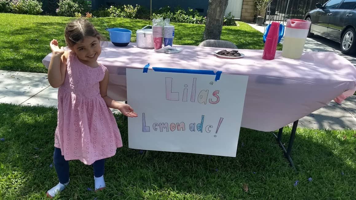 Child standing in front of the table of their lemonade stand smiling. 