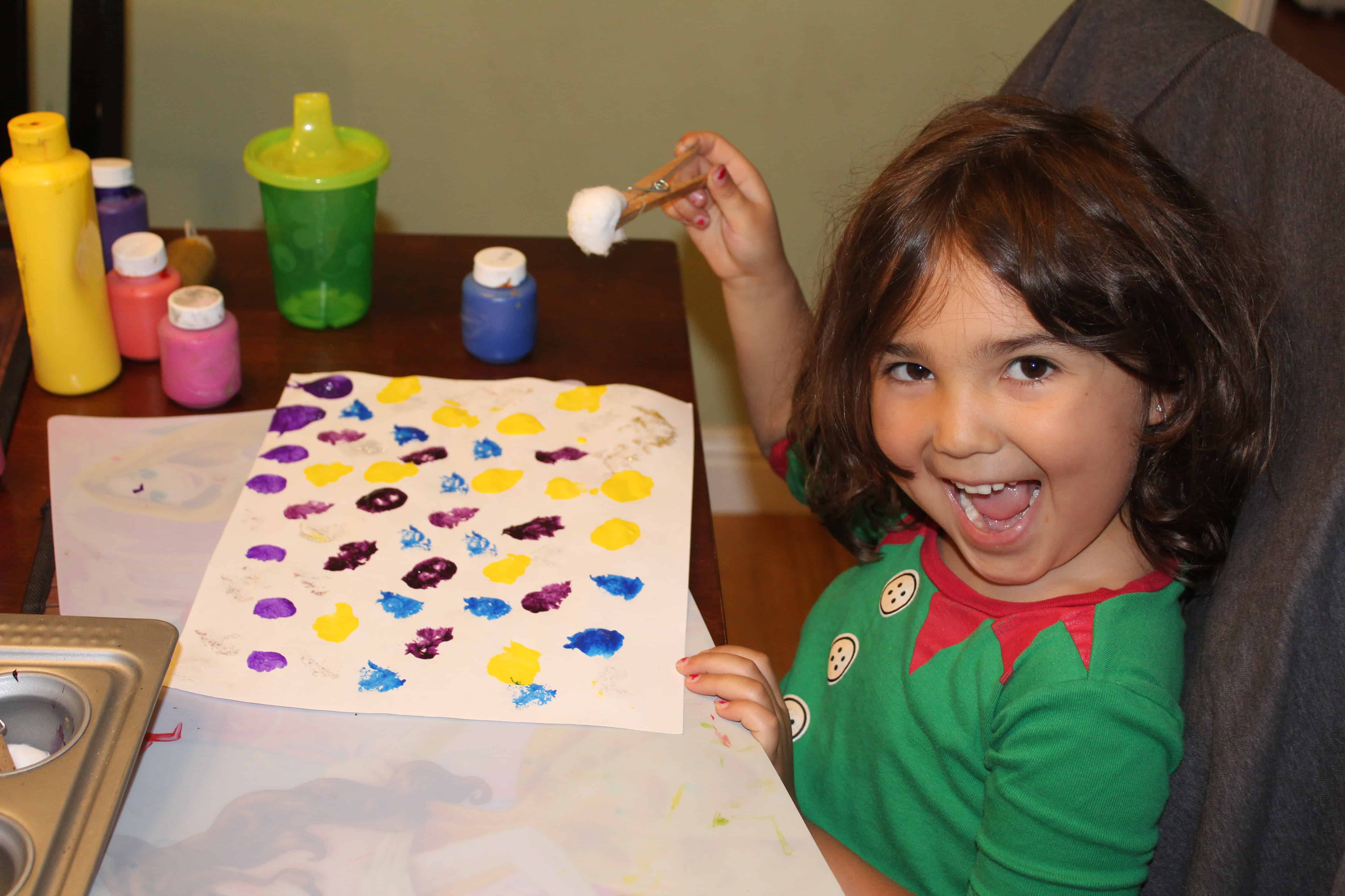 Young girl smiling painting with a clothespin cotton ball on a white sheet of paper