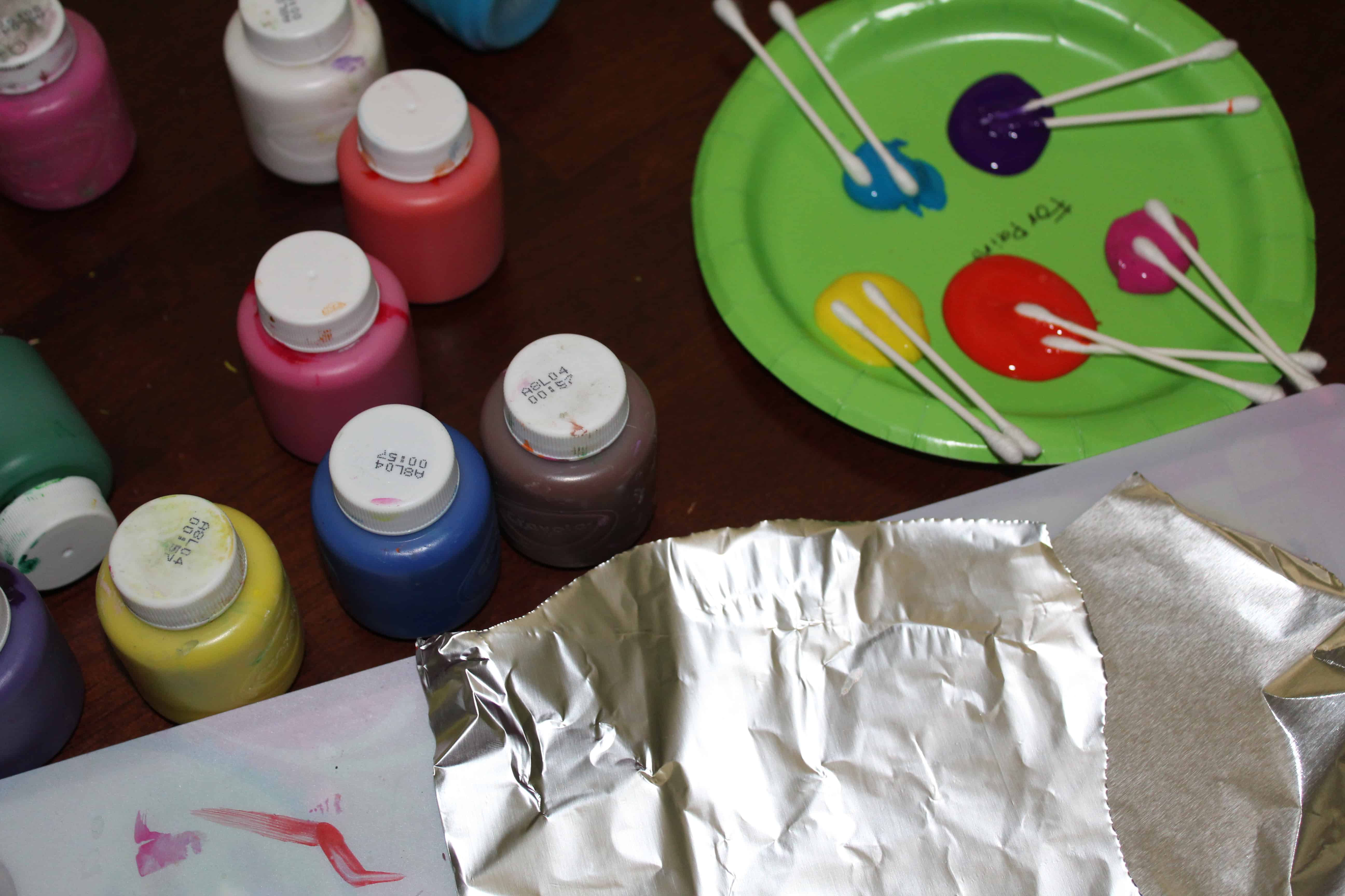 A paper plate with Acrylic paints on it. Containers of paint and a sheet of foil. 