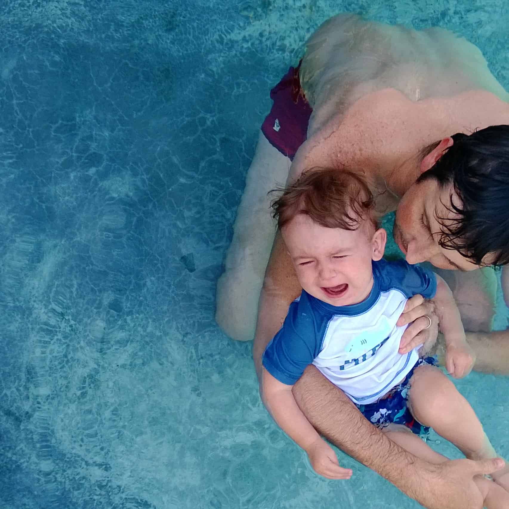 Toddler floating in the water while being held and crying. 