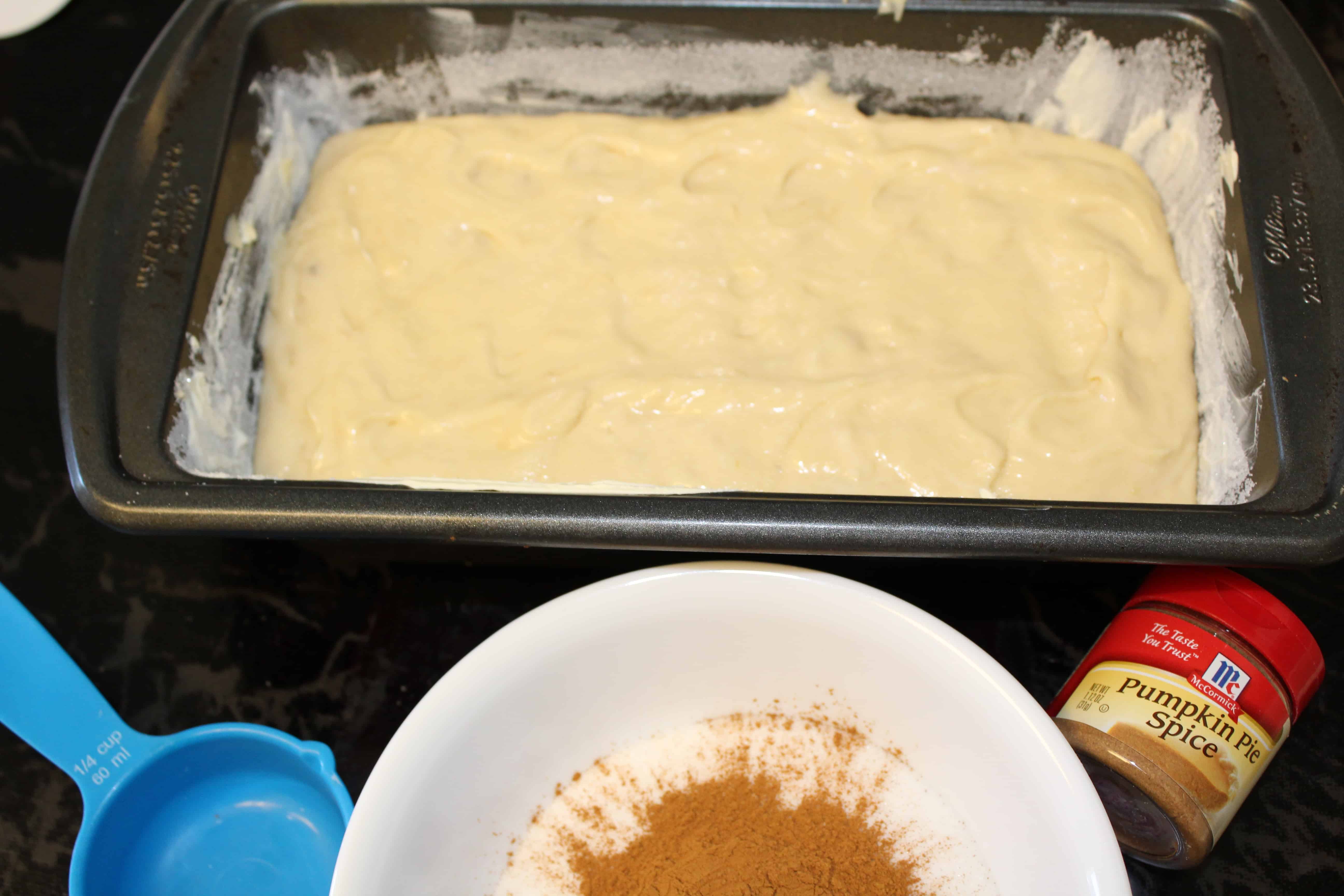 Batter poured evenly into a prepped bread pan next to a bowl of sugar and pumpkin spice.