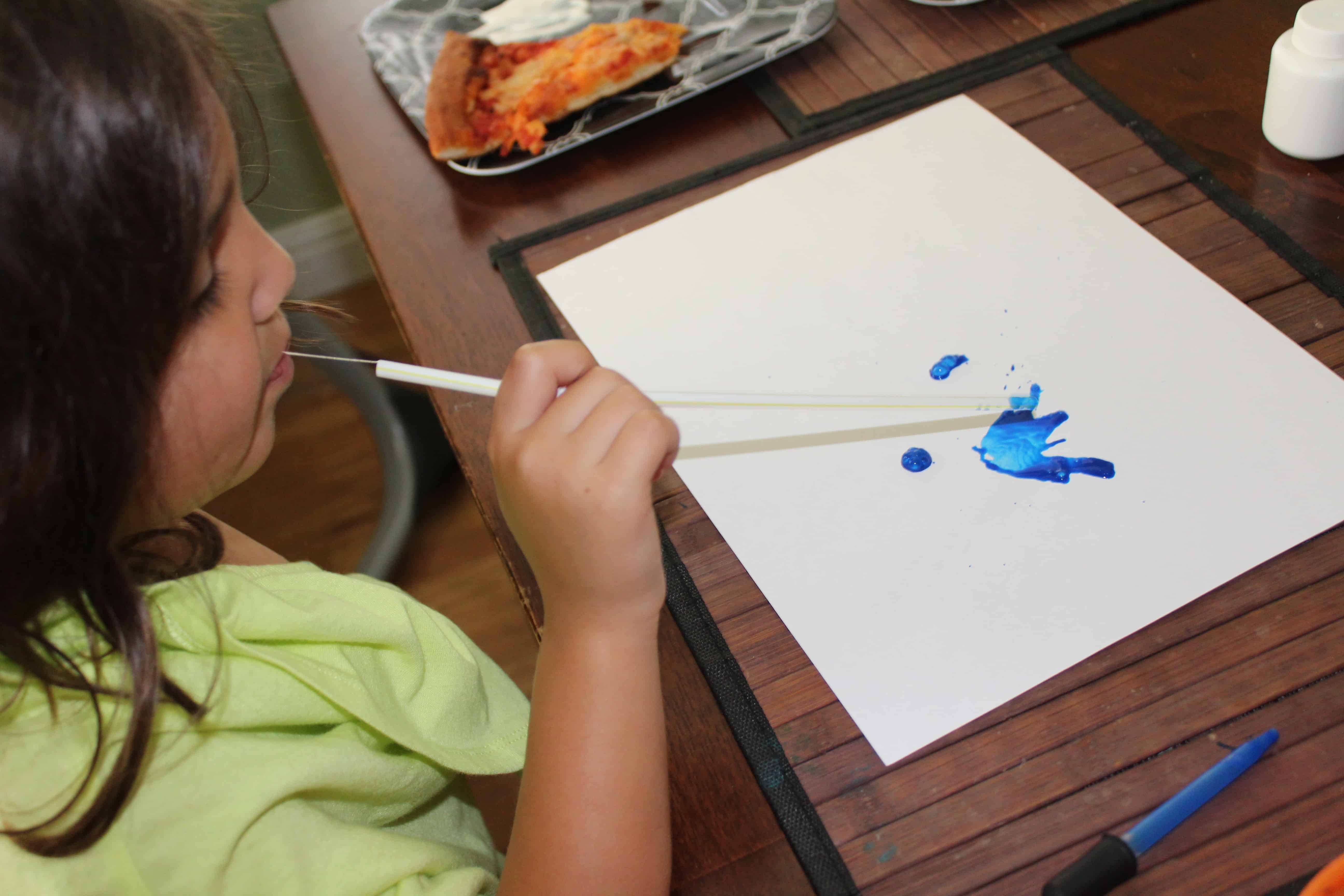 Little Girl using straw to blow paint patterns on a white sheet of paper. 