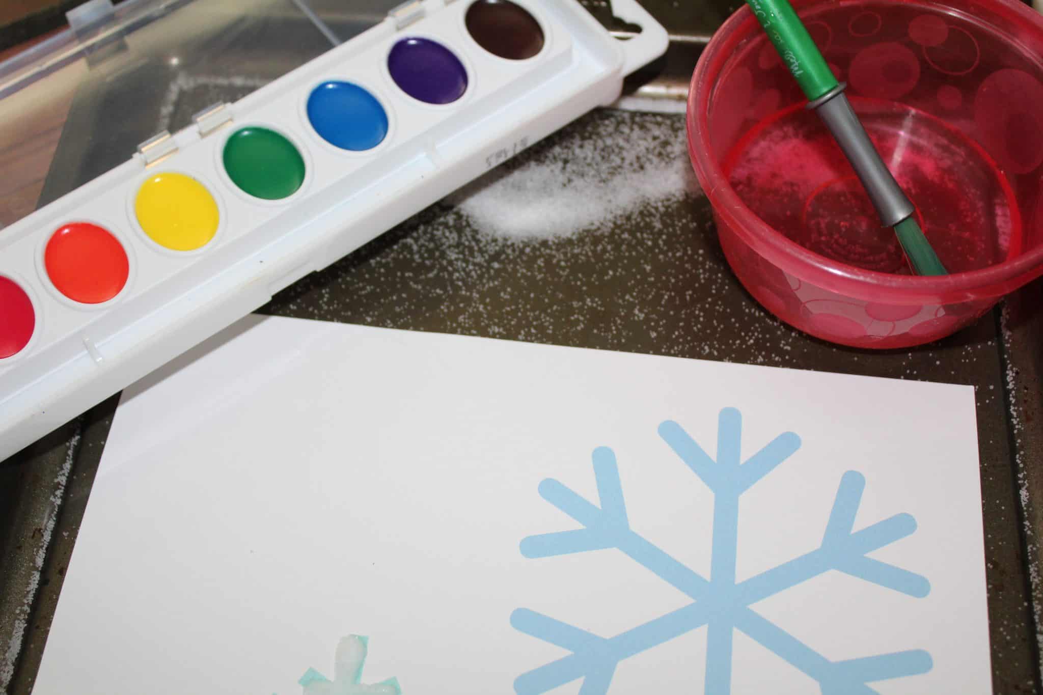 A pallet of water colors and a paint brush sitting next to a sheet with snowflakes templates