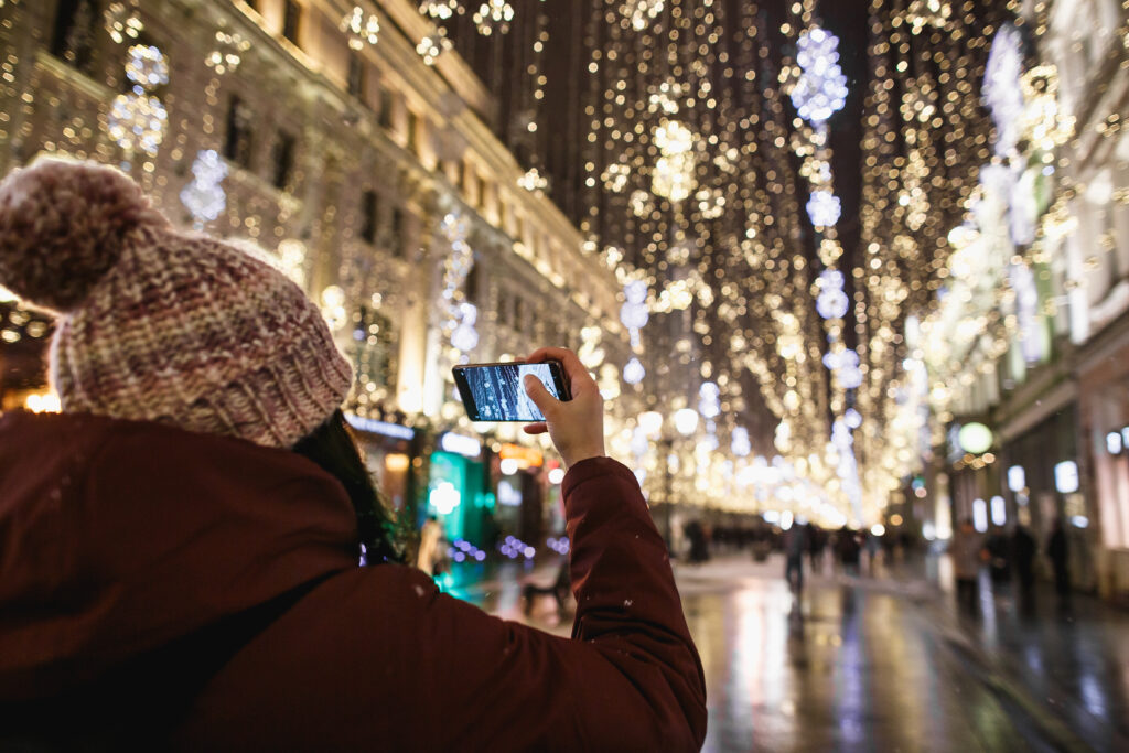 Outdoor night portrait of young fashionable woman in the winter hat with pompom taking photo by phone rear view. Magic snowfall effect. Night street illumination in Moscow.