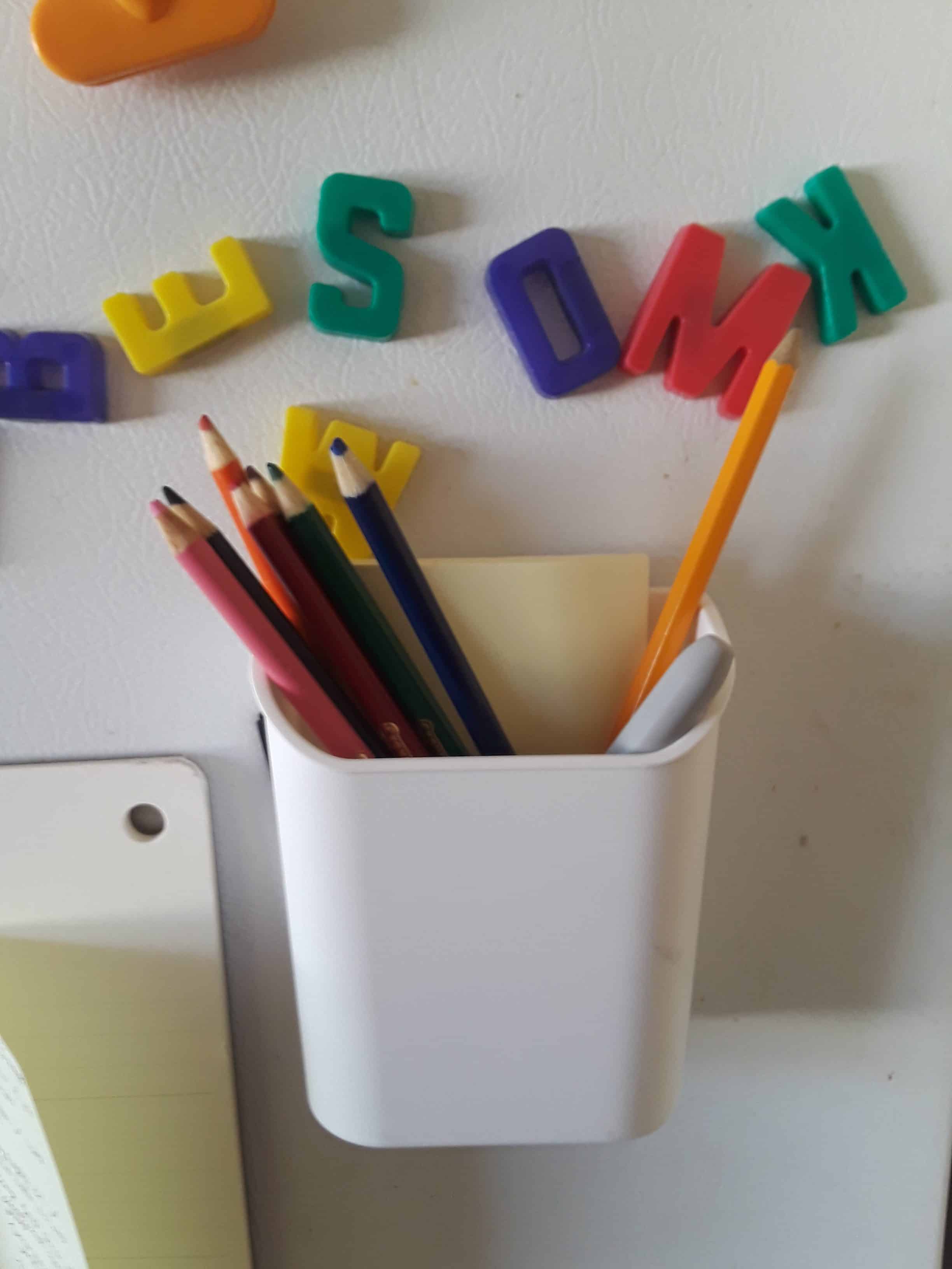 An array of of colored pencils in a magnetic cup holder with magnetic letters on the fridge door