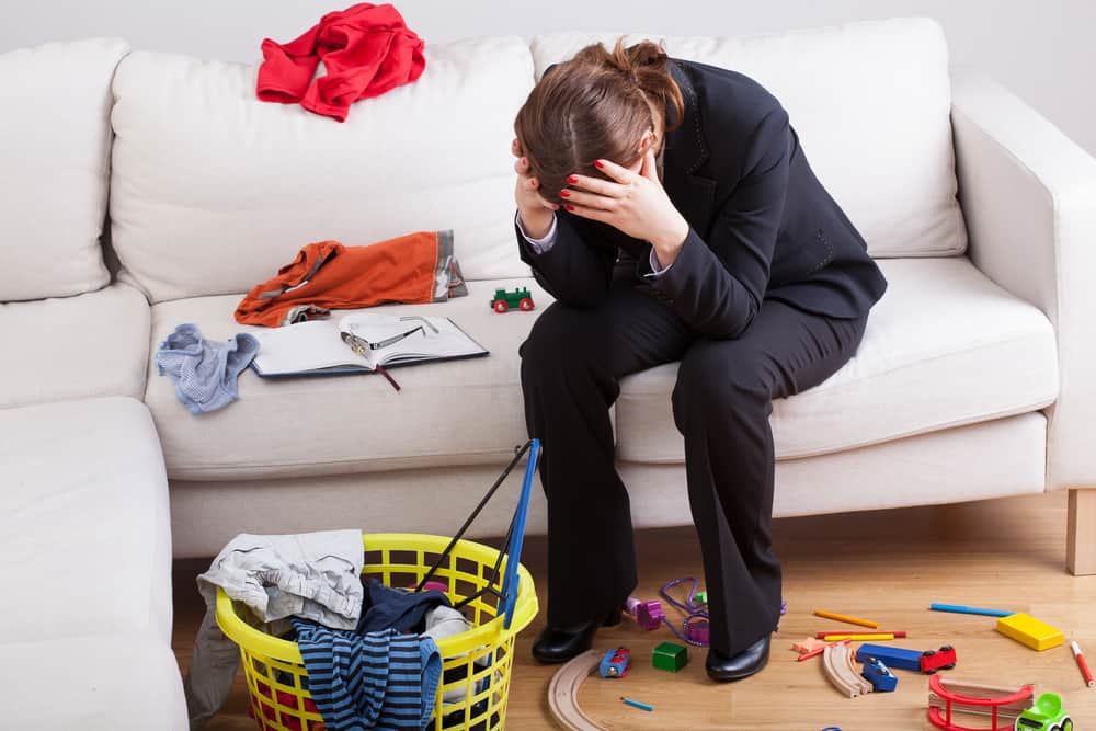 Frustrated woman with head in hands on a couch with clothes and toys spread around.