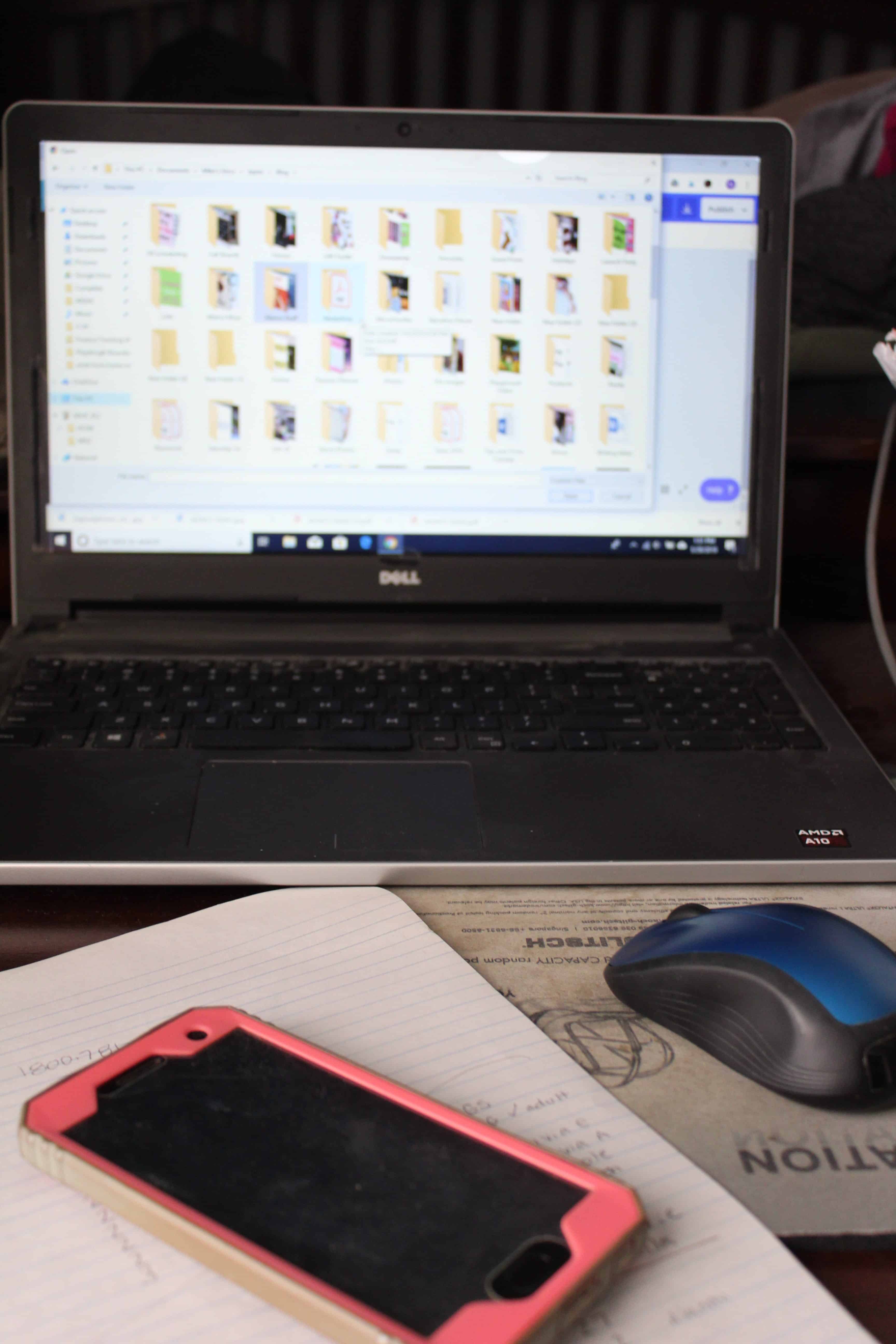 An open laptop screen with some filed folders open, a book and a cellphone along with a mouse on a desk. 