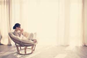 Young woman at home sitting on modern chair in front of window relaxing in her lliving room reading book and drinking coffee or tea