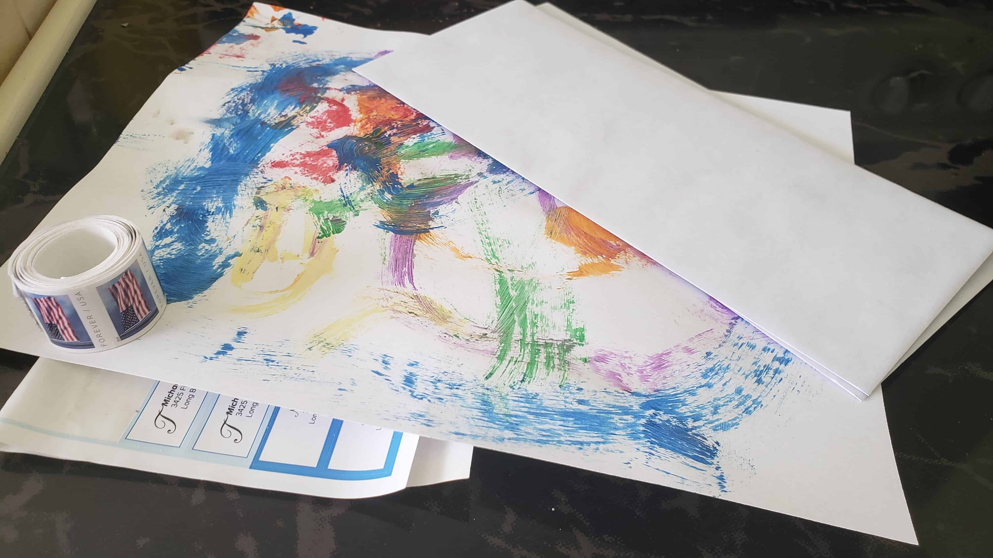 A roll of stamps, an envelope and a sheet of paper with painted artwork on it. 