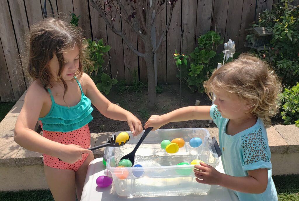 Two little girls using spoons to scoop plastic Easter eggs out of a plastic bin full of water