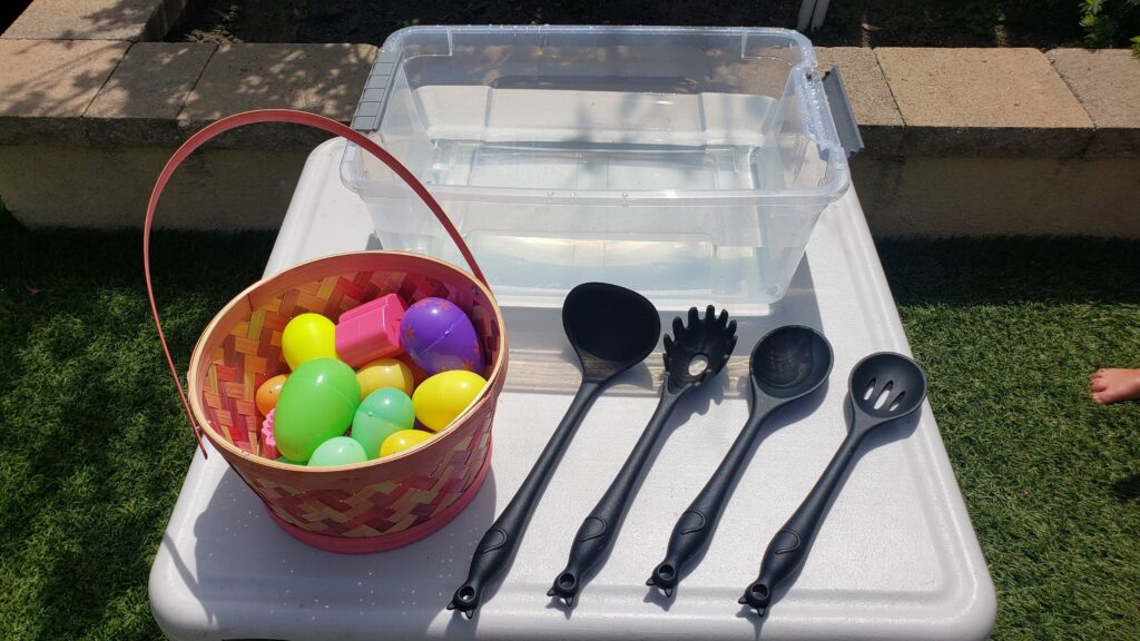 Plastic tub filled with water, plastic Easter eggs and four different styles of kitchen spoons